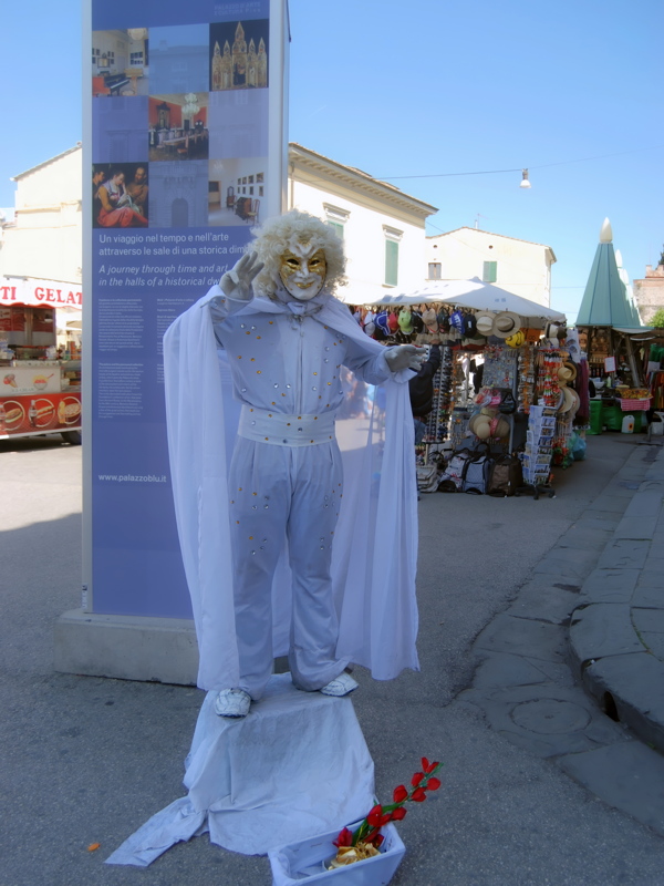 4 - A street performer acting as a living statue, dressed in a white costume and mask with a cape, posing on a small platform on a public square